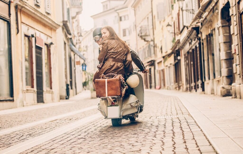 A couple riding a classic Italian moped through a narrow, picturesque alleyway in Italy, surrounded by historic buildings.