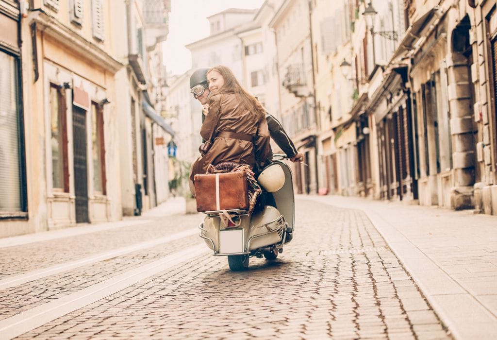 A couple riding a classic Italian moped through a narrow, picturesque alleyway in Italy, surrounded by historic buildings.