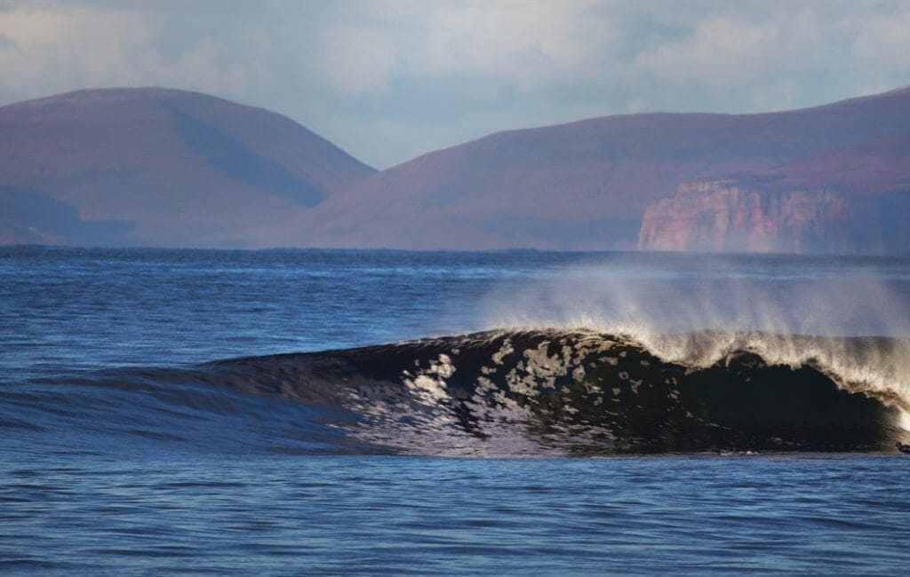 A surfer carves through a massive right-hander at Thurso East against a backdrop of the Scottish Highlands