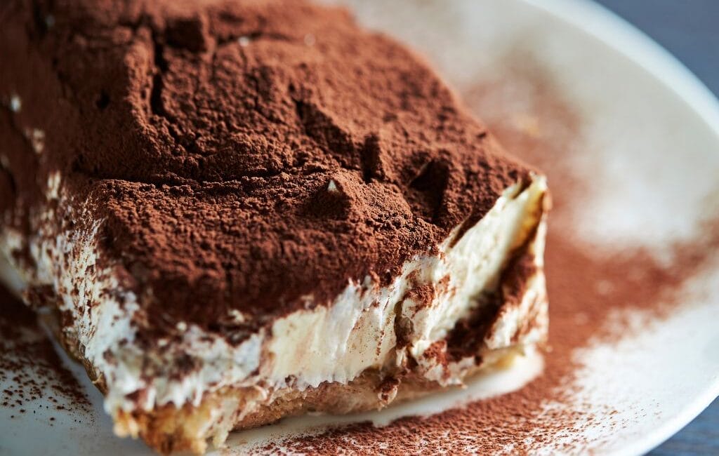 A perfectly layered slice of authentic Italian Tiramisu, topped with cocoa powder and dark chocolate shavings, served on a white plate