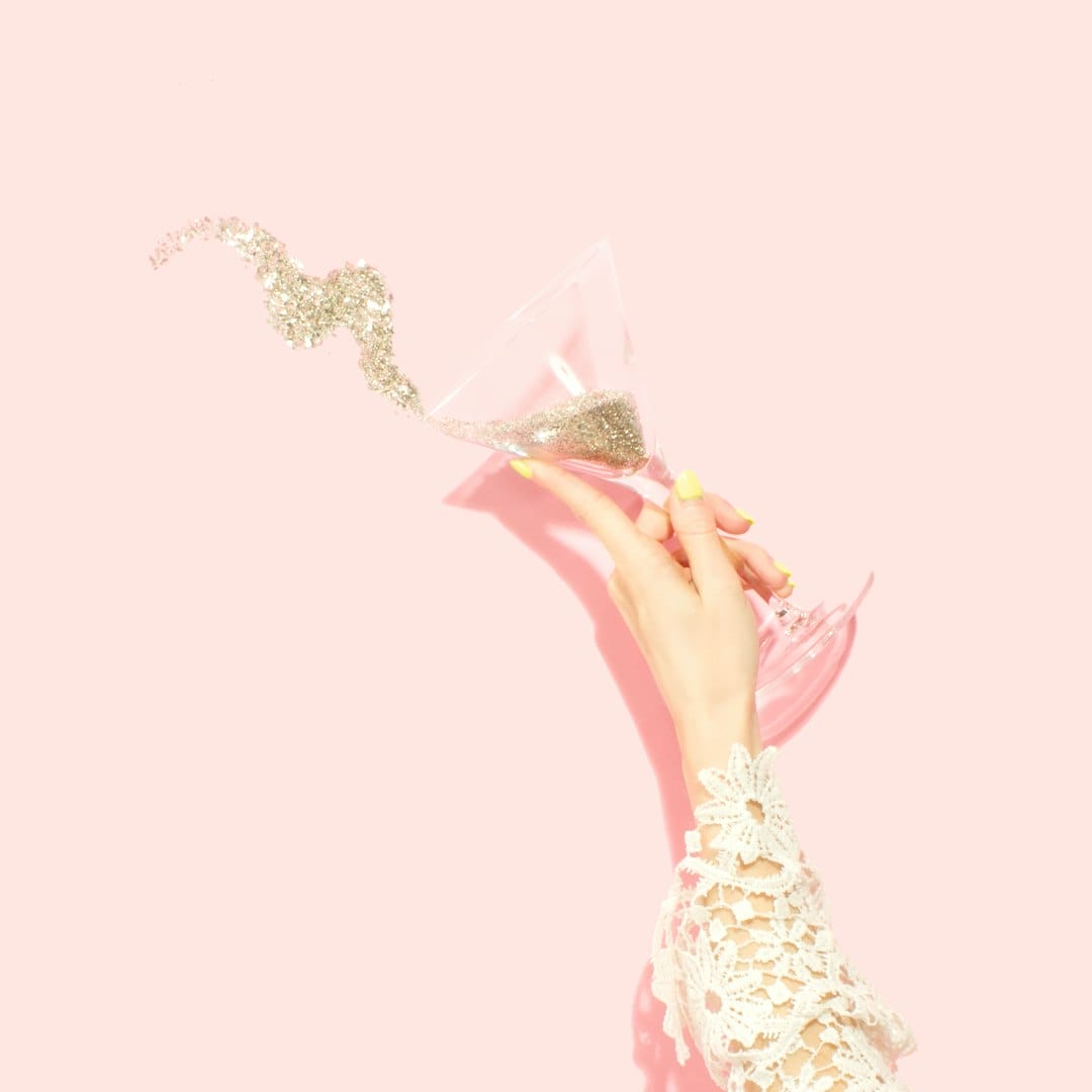 Hand holding a champagne glass with gold flowing out, set against a light pink background, symbolizing luxury and Après-Ski in St. Anton, Austria.